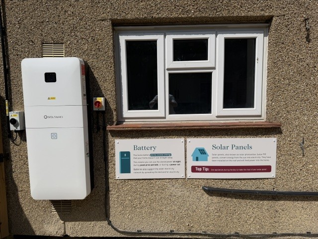 Home battery on the outside wall of the house, next to information signs about the battery and solar panels