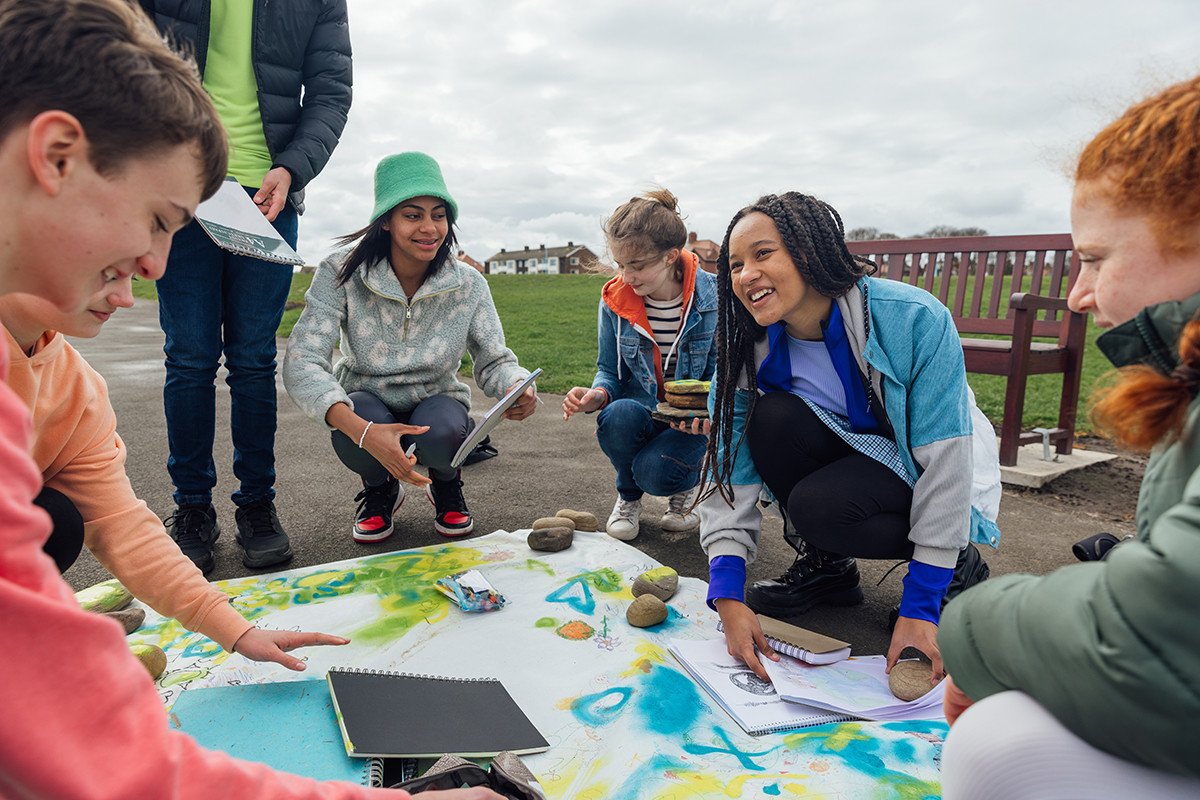 Group of young people crouching around a large painted map on the ground outdoors, planning together