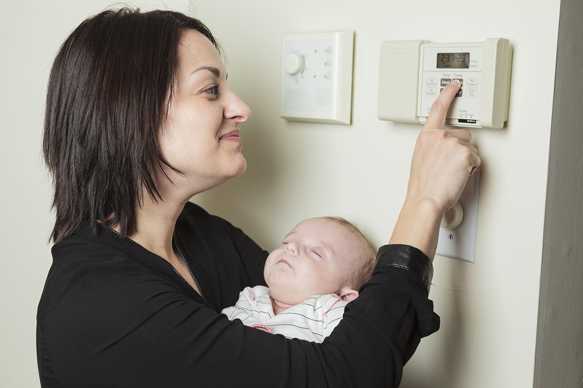 Woman holding a sleeping baby while adjusting the heating thermostat on the wall